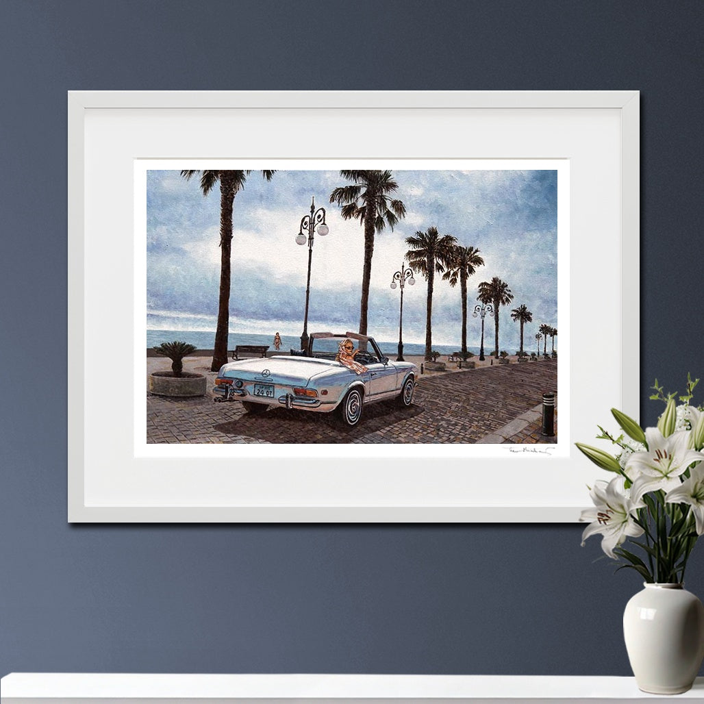 A framed art print depicting a vintage convertible car on a palm-lined boardwalk by the ocean, displayed on a white surface against a dark blue-gray wall with white lilies in a vase.